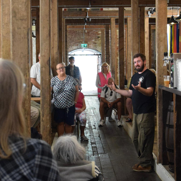 A man in a black t-shirt and green cargo pants stands in a rustic brewery or workshop hallway, gesturing to a small group of people who are listening. The setting features exposed wooden beams, brick walls, and a wooden floor.