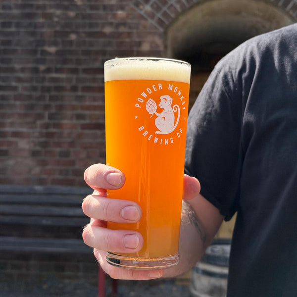A person holding a pint glass of orange beer with a white foam head. The glass has a white printed logo that reads 'Powder Monkey Brewing Co' with a monkey illustration.